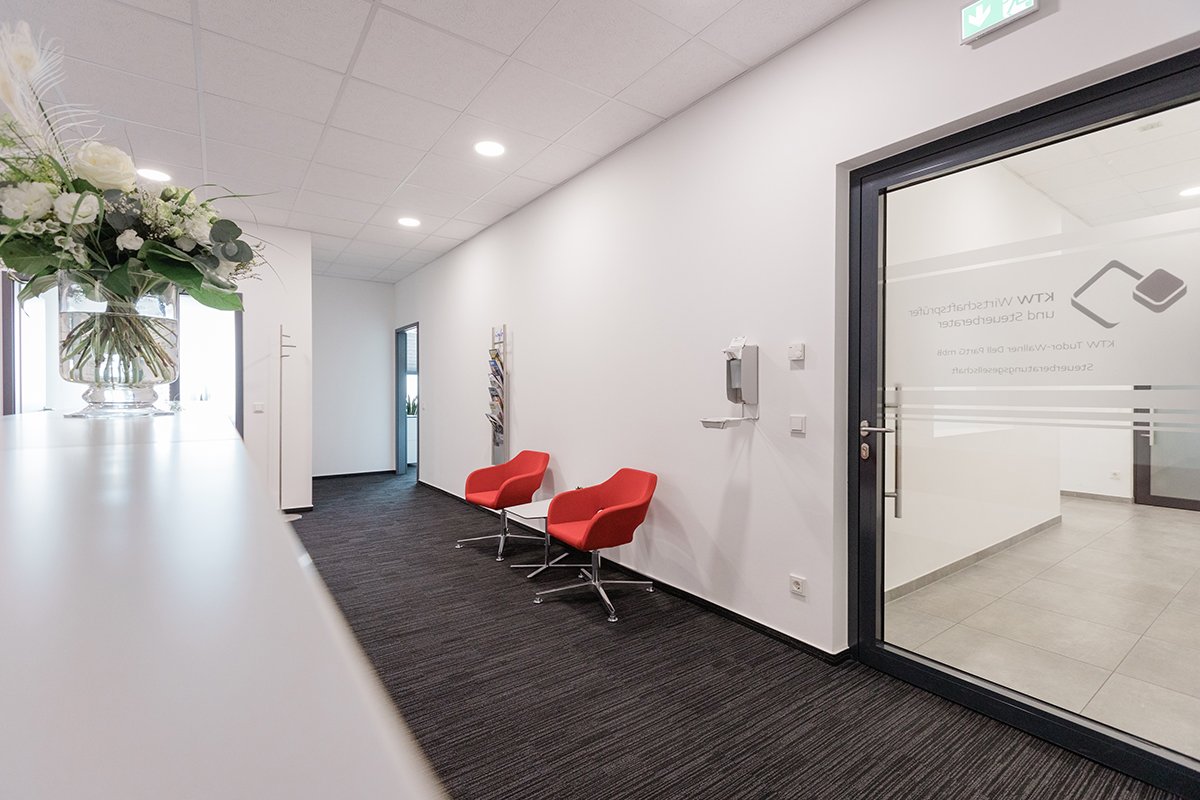 Moderner Kanzlei-Büroflur mit zwei roten Stühlen, Glastür und Blumengesteck auf dem Tresen.