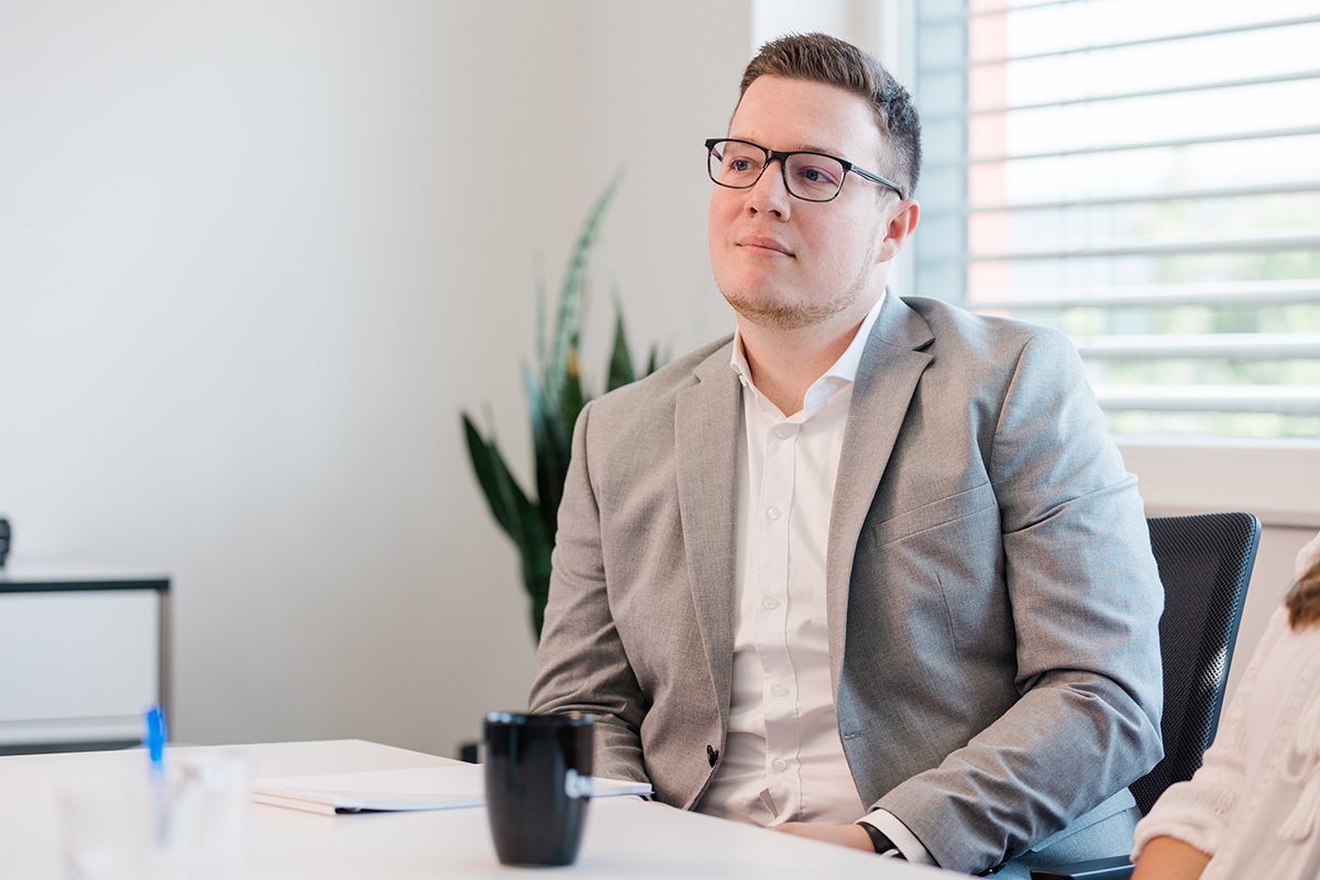 Steuerberater in grauem Anzug am Schreibtisch mit Notizbuch und KTW-Tasse, wirkt aufmerksam.