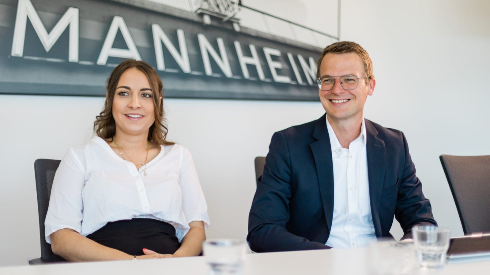 Zwei Personen in Geschäftskleidung sitzen lächelnd an einem Tisch, hinter dem ein Schild der KTW-Kanzlei hängt. Zwei Personen in Geschäftskleidung sitzen lächelnd an einem Tisch, hinter dem ein Schild der KTW-Kanzlei hängt.