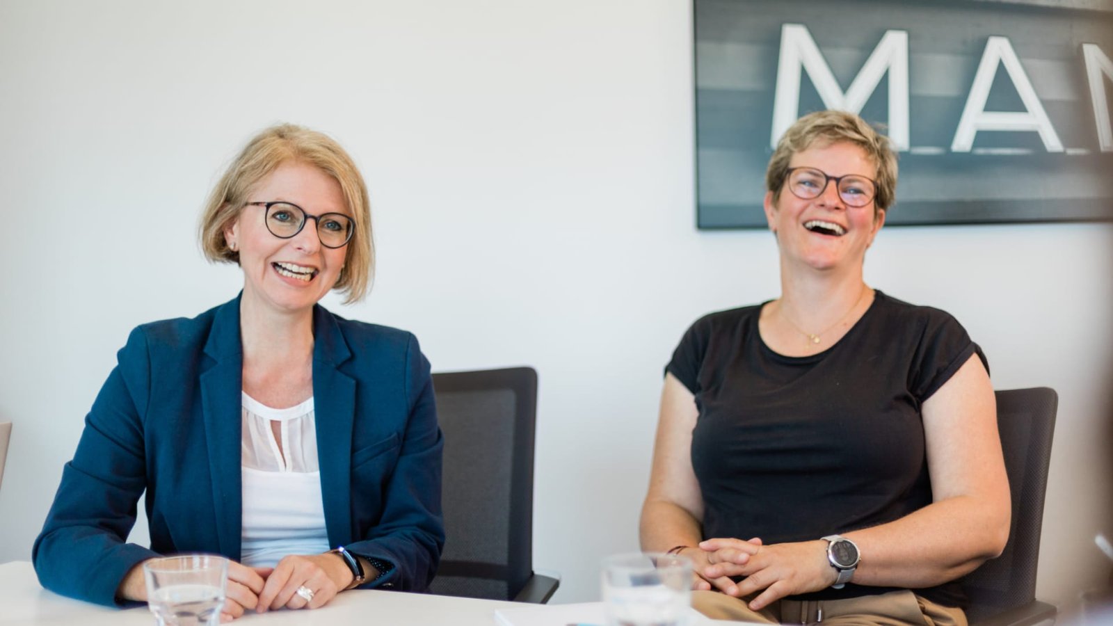 Zwei Frauen lächeln an einem Tisch in einem hellen, modernen Büro der Kanzlei. Zwei Frauen lächeln an einem Tisch in einem hellen, modernen Büro der Kanzlei.